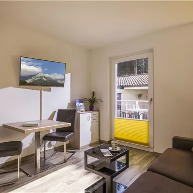 A bright living space with a modern sofa, a table, and chairs. A television and a window with yellow blinds complete the ambiance.