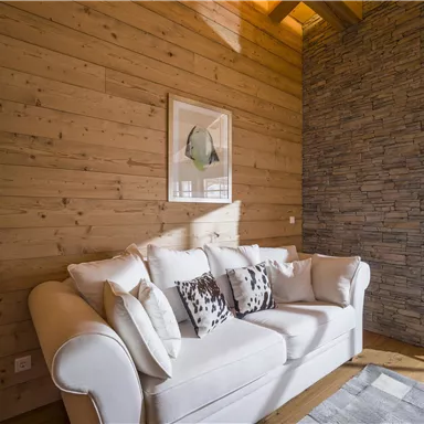 Ein gemütliches Wohnzimmer mit einem weißen Sofa und dekorativen Kissen. Die Wände sind aus Holz, und ein Steingemäuer ergänzt die warme Atmosphäre.