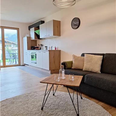 Ein modernes Wohnzimmer mit einem grauen Sofa und einem Holztisch. Eine offene Küche ist im Hintergrund sichtbar, und große Fenster sorgen für viel Tageslicht.