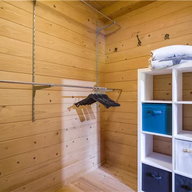 Ein geräumiger Kleiderschrank mit Holzverkleidung. Auf der linken Seite hängen Kleiderbügel, rechts steht ein Regal mit bunten Aufbewahrungsboxen.