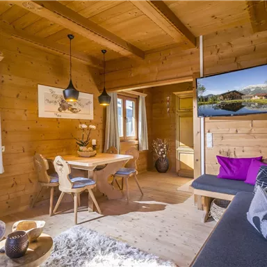 Ein gemütlicher Wohnraum in einer Holz-Hütte mit einem Esstisch und einem Fernseher. Große Fenster lassen viel Licht herein und schaffen eine einladende Atmosphäre.
