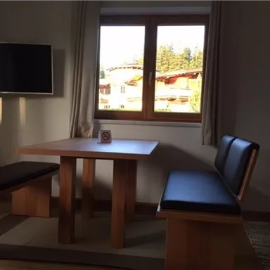 Ein helles Zimmer mit einem Tisch und Bank in Holzoptik. Im Hintergrund sind Fenster mit Blick auf ein schönes Landschaftsbild.