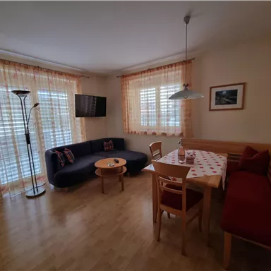 Ein helles Wohnzimmer mit einem dunklen Sofa und einem Esstisch. Große Fenster mit hellen Vorhängen lassen viel Licht herein.