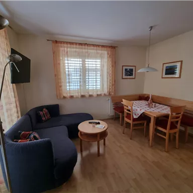 Ein gemütliches Wohnzimmer mit einer dunkelblauen Couch und einem Holztisch. Der Raum ist hell, mit Fenstern, die Tageslicht hereinlassen, und einer Essecke.