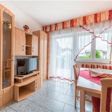 A cozy living space with a dining area and a television. Large windows with bright curtains provide plenty of light.