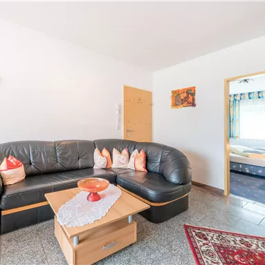 A cozy living room with a black sofa and a wooden table. In the background, a bedroom and some decorative elements can be seen.