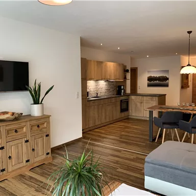 A modern living room with light wood furniture and an open kitchen. The decor is cozy and inviting, with plants and warm lighting.