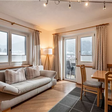 A cozy living kitchen with a bright sofa and a dining table. Large windows provide a view of the surroundings.