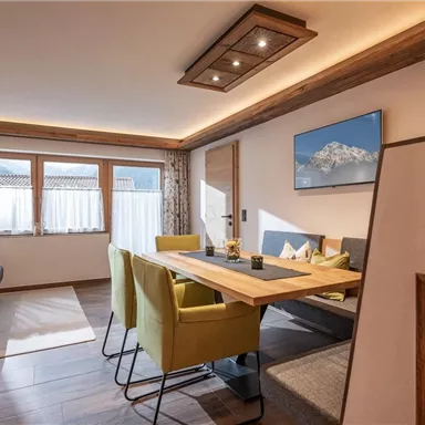 A modern living room with a dining table and green chairs. The windows provide daylight and a view of a mountain landscape.