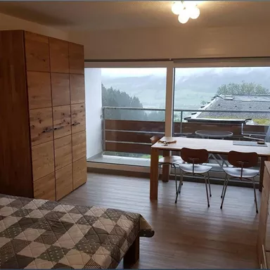 A bright room with a bed and a wooden wardrobe. The balcony offers a view of the mountains and the landscape.
