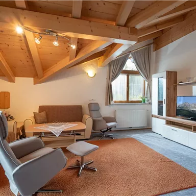 A modern living room with a wooden ceiling and cozy furniture. A TV and large windows provide plenty of light and an inviting atmosphere.