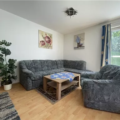 Ein gemütliches Wohnzimmer mit einer grauen Couchgarnitur und einem Tisch. Grosses Fenster mit Blick ins Grüne und dekorative Bilder an der Wand.