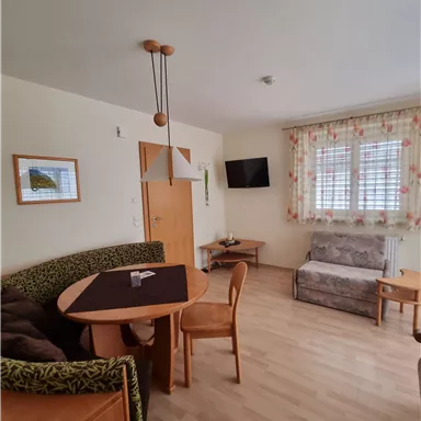 Ein helles Wohnzimmer mit einem Esstisch und Stühlen. Es gibt eine Couch und einen Fernseher an der Wand, sowie Fenster mit Vorhängen.