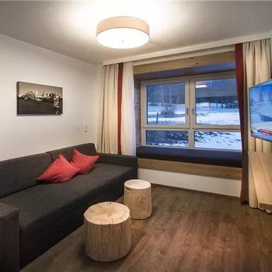 A modern living room with a gray sofa and red cushions. There is a television and a view of a snow-covered landscape through the large window.