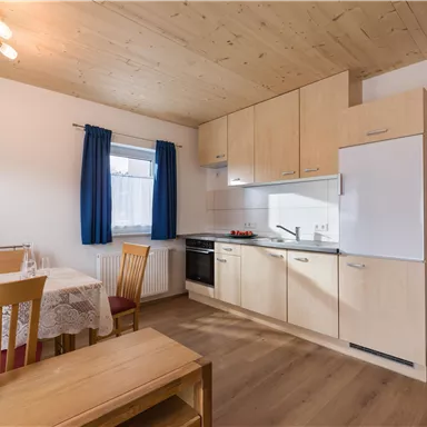 A modern kitchen with wood decor and light furniture. The room has a table with chairs and a window with blue curtains.