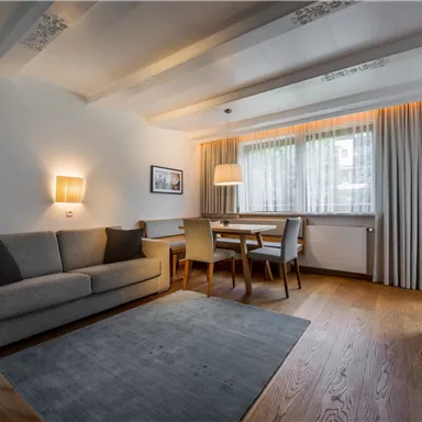 A cozy living room with a gray sofa, a dining table, and chairs. Generous windows with curtains create a bright ambiance.