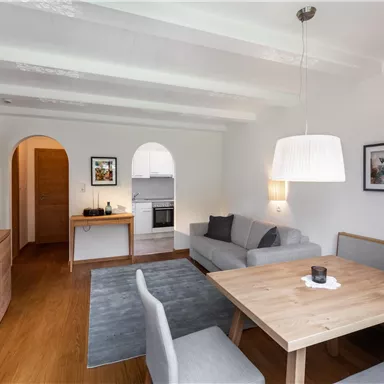 A modern living room with bright furniture and a large table. There is a cozy seating area and an open kitchen in the background.