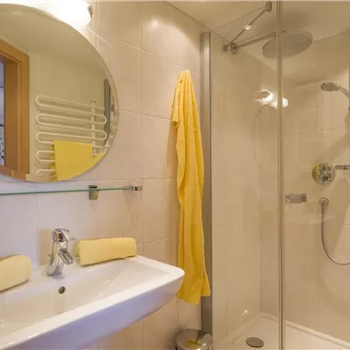 Ein modernes Badezimmer mit einem runden Spiegel und einer gläsernen Duschkabine. Gelbe Handtücher und eine sanfte Beleuchtung schaffen eine einladende Atmosphäre.