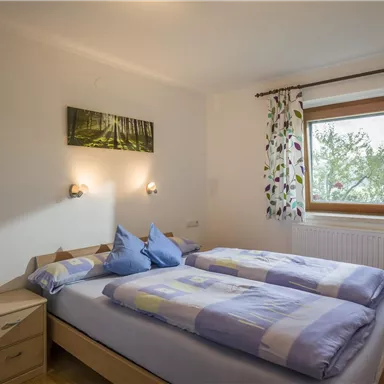 Ein gemütliches Schlafzimmer mit zwei Betten und bunten Kissen. Ein Fenster bietet einen Blick ins Freie, und dekorative Vorhänge ergänzen den Raum.