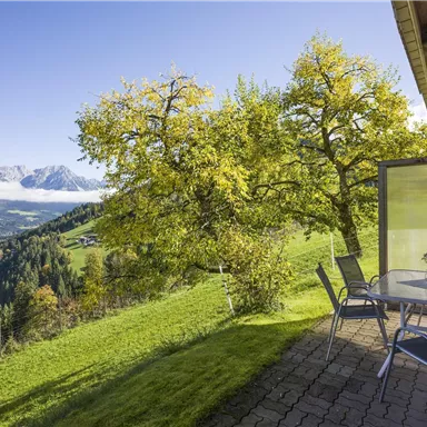 Ein schöner Ausblick auf die Berge mit einem grünen Hügel und bunten Bäumen. Ein Tisch mit Stühlen steht auf einer Terrasse.