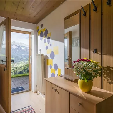 Ein einladender Flur mit Holzvertäfelung und einem offenen Eingang. Auf einem Möbelstück steht eine Vase mit bunten Blumen und im Hintergrund sind die Berge sichtbar.