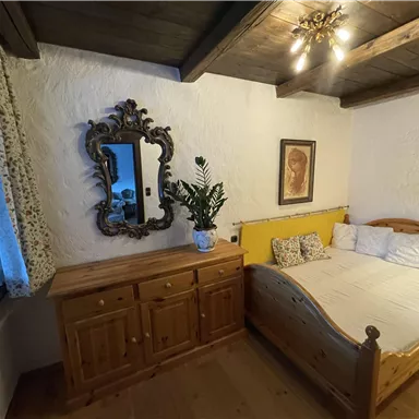 A cozy bedroom with a wooden bed and a dresser. A beautiful mirror hangs on the wall, and a plant stands on the dresser.