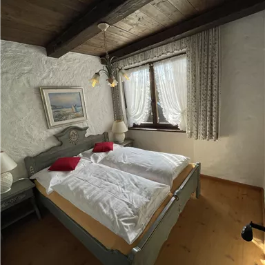 A cozy bedroom with a double bed and red pillows. The room has wooden ceilings and large windows with curtains.