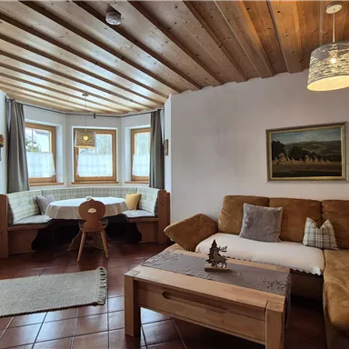 A cozy living room with a wooden beam ceiling and large windows. The room is furnished with a comfortable couch, a table, and rustic decorations.
