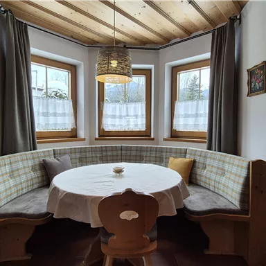 A cozy dining area with a round table and a corner bench. Large windows let in plenty of daylight.