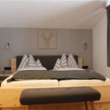 Ein gemütliches Schlafzimmer mit einem großen Doppelbett und modernen Holzmöbeln. An der Wand hängt ein Bild eines Hirschkopfes, und das Zimmer hat große Fenster mit Vorhängen.