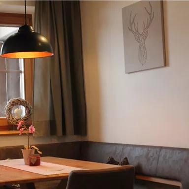 Ein gemütlicher Essbereich mit einer Holzbank und einem Tisch. Eine moderne Lampe hängt über dem Tisch, und ein antler-artiges Kunstwerk schmückt die Wand.