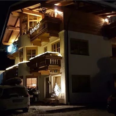 Ein gemütliches Haus im Winter, beleuchtet mit Weihnachtslichtern. Schnee liegt auf dem Boden und Autos stehen davor.