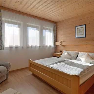 A cozy bedroom with a double bed and an armchair. Large windows provide plenty of daylight in a modern wooden design.