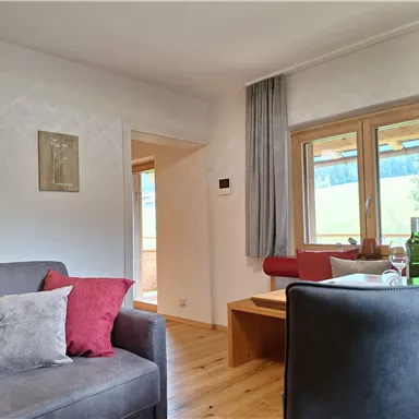 A cozy living room with a gray sofa and red cushions. Large windows offer a view of the green landscape outside.