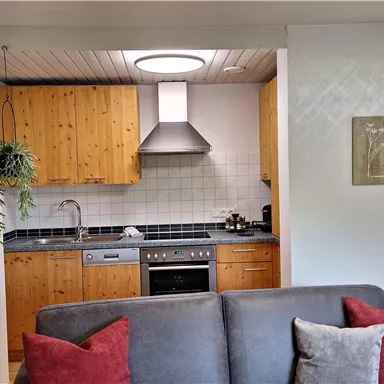 A modern kitchen with wooden furniture and a stove. In the foreground, there is a gray sofa with red cushions.