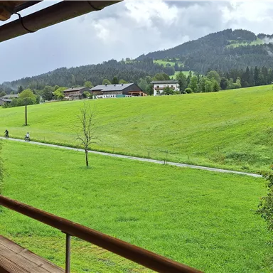 A green meadow with gentle hills and houses in the background. A winding path stretches through the landscape.