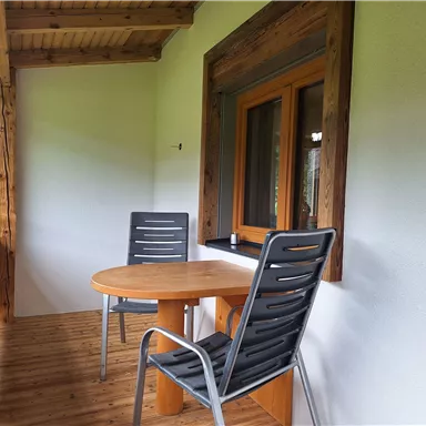 A small balcony with a round table and two chairs. The walls are painted light and there is a window with wooden elements.