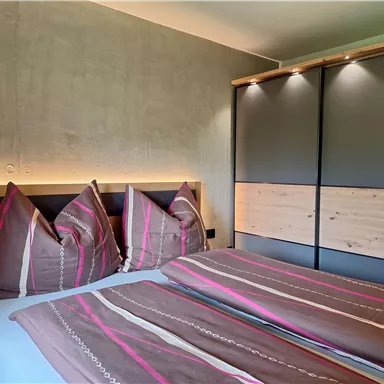 A cozy bedroom with a double bed and modern wooden furniture. The walls are plain, and the lighting creates a pleasant atmosphere.