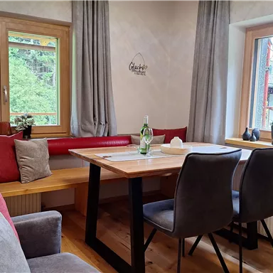 A cozy dining area with a wood-colored table and modern chairs. The windows offer a view of the green surroundings and let in plenty of light.