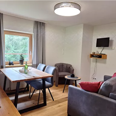 A modern living room with a wooden table and cozy armchairs. Large windows provide plenty of daylight and a view of the greenery.