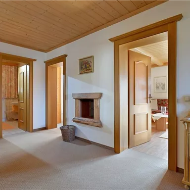 A bright hallway with wooden walls and doors. It leads to several rooms and has a fireplace as well as a small dresser.