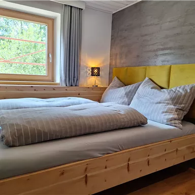 A cozy bedroom with a large bed and yellow headboard. Windows overlooking nature and gentle curtains.