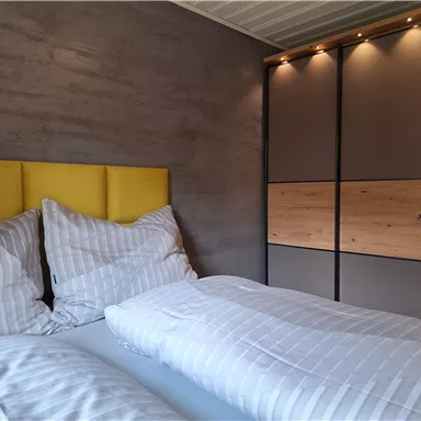 A modern bedroom with a yellow headboard and two striped bedcovers. The room is designed with a wardrobe and soft lighting.