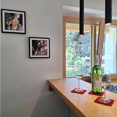 A cozy bar with a wooden bar and two wine glasses. Decorative pictures hang on the wall, and large windows let in plenty of light.