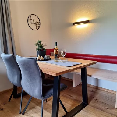 A cozy dining area with a wooden table, modern chairs, and a bench. On the table, there is a bottle of wine and glasses, while soft light illuminates the room.