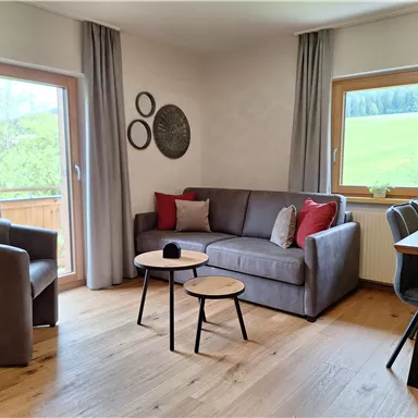 A cozy living area with a gray sofa, a small table, and a dining area. Large windows offer a view of the greenery.