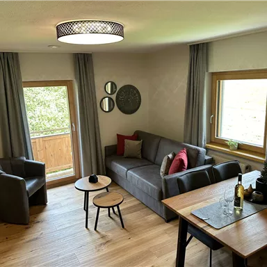 A modern living room with a gray sofa and a wooden table. Large windows allow plenty of daylight and offer a view of the outdoors.