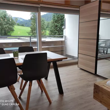 Ein modernes Zimmer mit einem Esstisch und Stühlen. Durch große Fenster ist eine grüne Landschaft im Hintergrund sichtbar.