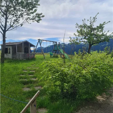 Ein Spielplatz mit einer Schaukel und einer kleinen Hütte in einer grünen Umgebung. Im Hintergrund sind Berge und ein blauer Himmel sichtbar.