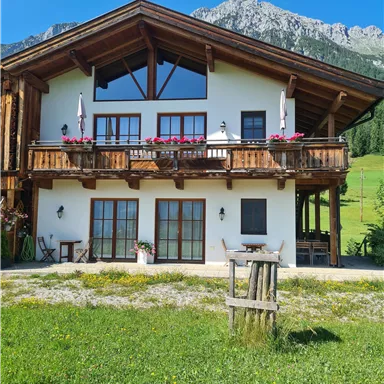 Ein schönes, zweistöckiges Holzhaus mit einem großen Balkon. Im Hintergrund sind grüne Wiesen und Berge zu sehen.